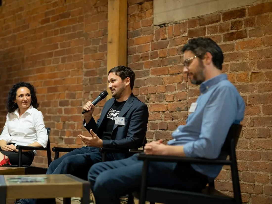 Three panellists seated on a stage against a brick wall, one of them speaking into a microphone.