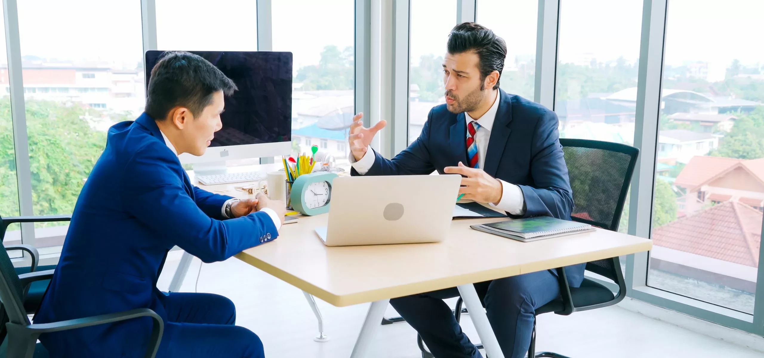 Two people talking across a meeting table with a laptop between them.