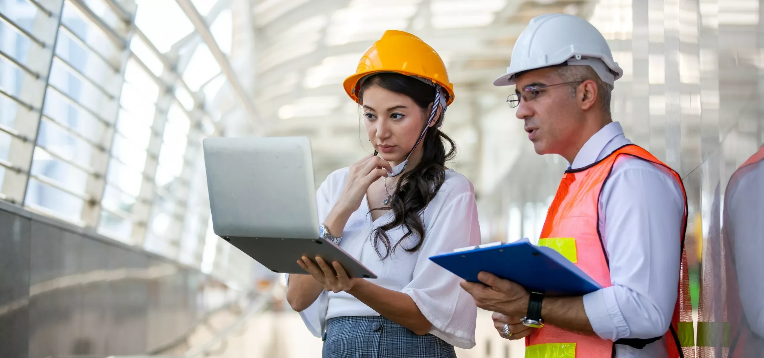 An engineer in a hard hat with a laptop, beside a colleague in a hi-vis vest holding a clipboard.
